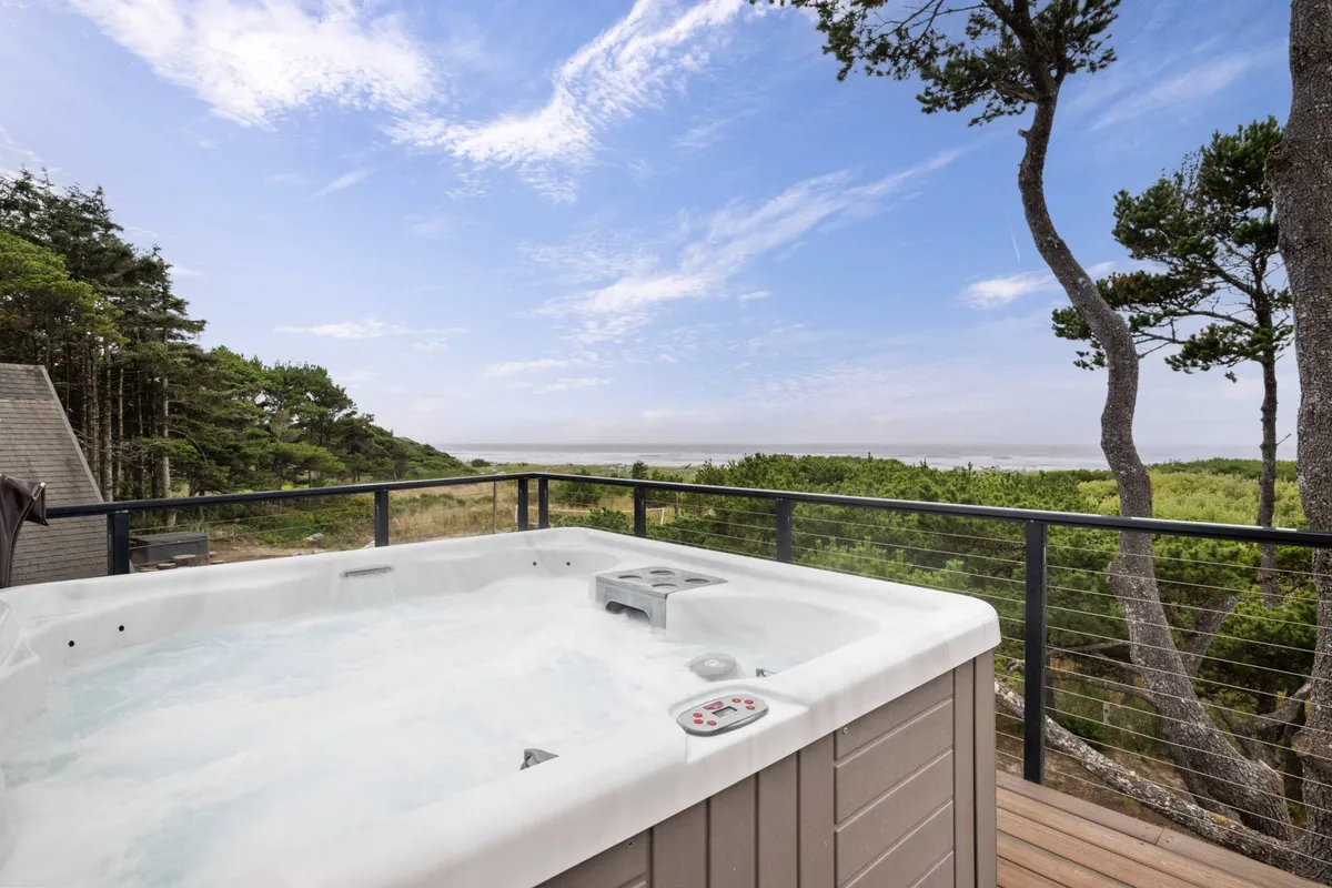A tranquil exterior view featuring a spacious deck with a six-person hot tub in the foreground, surrounded by lush trees and offering a breathtaking panorama of the ocean. The arrangement is perfect for relaxing or socializing while soaking up the coastal scenery.