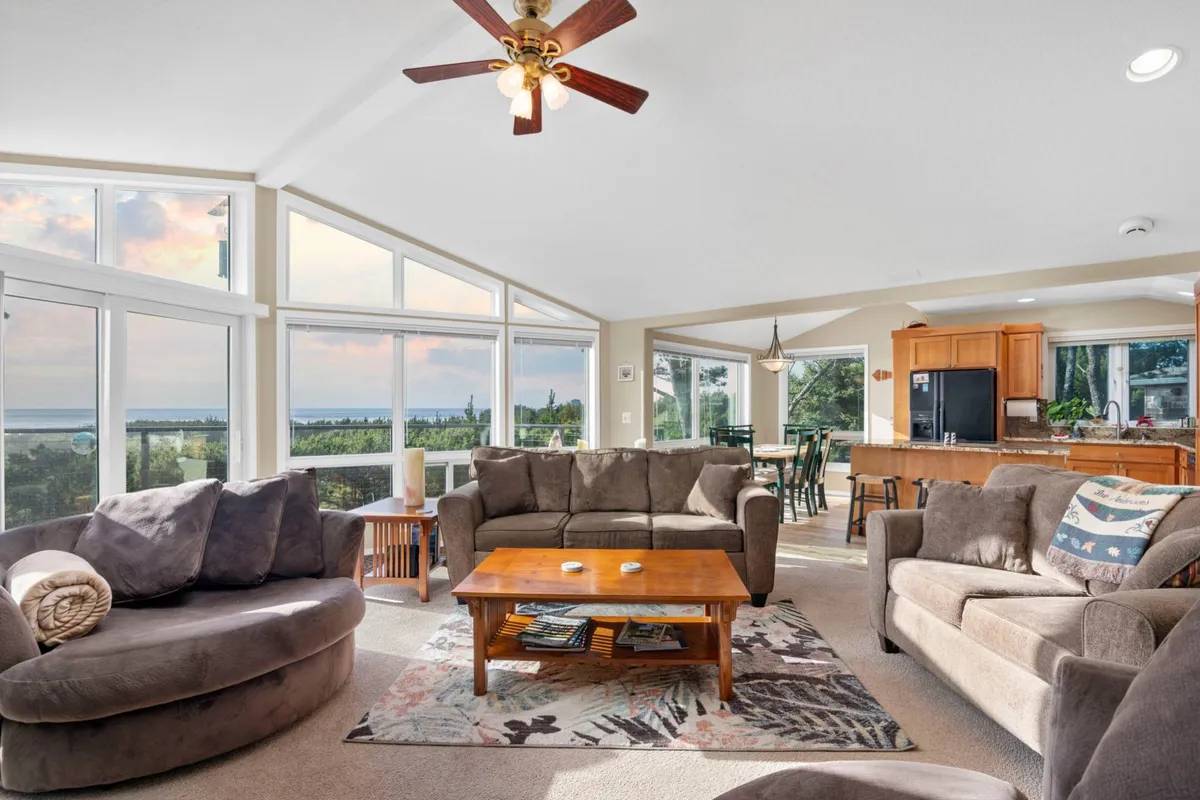 A cozy living room with large windows framing sweeping ocean views. Plush sofas and barrel chairs are arranged around a rustic wooden coffee table, creating an inviting space for group gatherings. The warm light from the sunset floods the room, and a flat-screen TV is visible, suggesting entertainment options.
