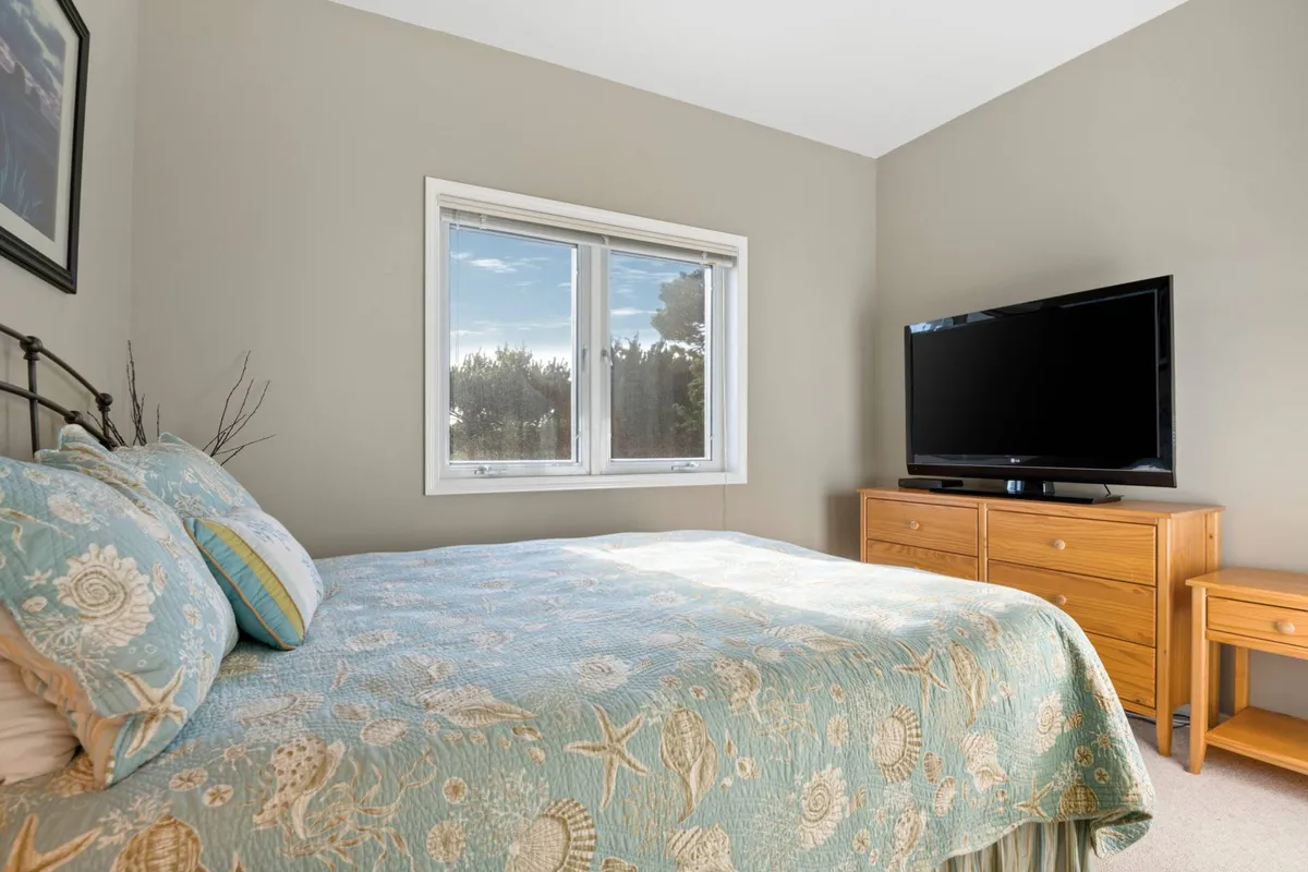 A serene bedroom with a bed covered in a blue patterned quilt, a large window letting in plenty of natural light, and a television on the dresser. The atmosphere is peaceful and cozy, ideal for unwinding at the end of the day or enjoying a private movie night.
