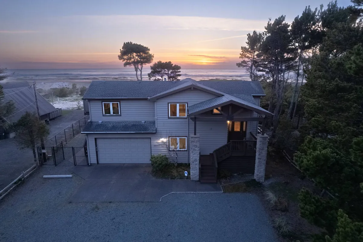 The hero image showcases a beautiful single-family beachfront house framed by lush coastal trees, with a wide porch facing the Pacific Ocean. The scene is bathed in warm, golden light from a stunning sunset, emphasizing the home's oceanfront location and tranquil surroundings. The setting invites relaxation, outdoor gatherings, and connection with nature.