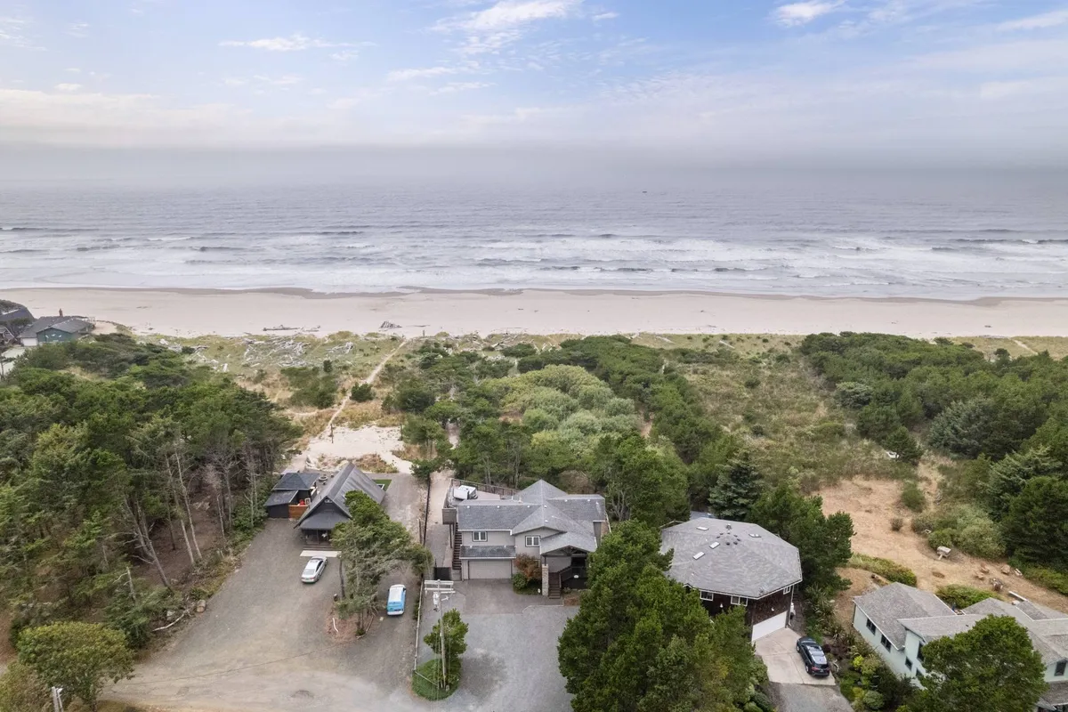 A sweeping view of the Oregon coastline with a sandy beach, lush trees, and the ocean extending to the horizon. Several houses sit nestled among the greenery, with the beach easily accessible for walks and playtime. The open, natural setting invites exploration and outdoor adventure.