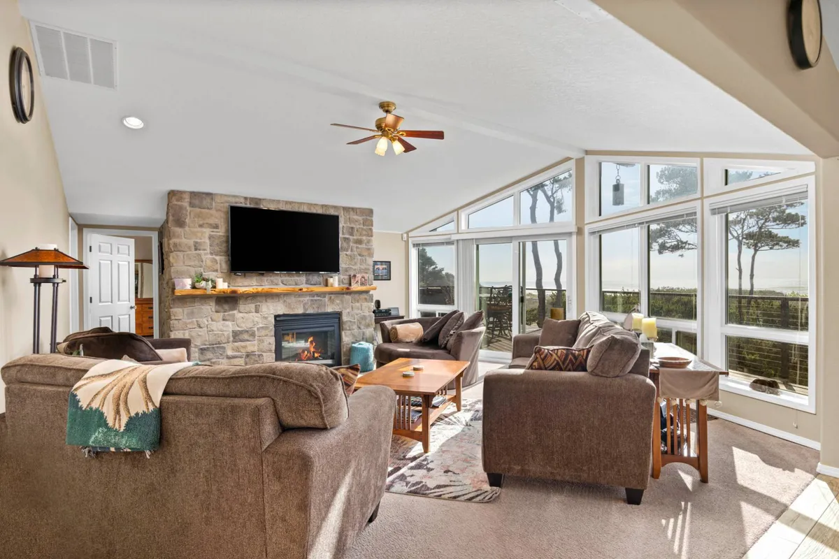 This image features a spacious, comfortable family room with a stone fireplace, large windows letting in natural light, and cozy seating arranged around a coffee table. The room is inviting and ideal for relaxing, watching TV, or spending quality time together. The view outside the windows hints at the natural surroundings, while the fireplace adds to the warmth and hominess of the space.