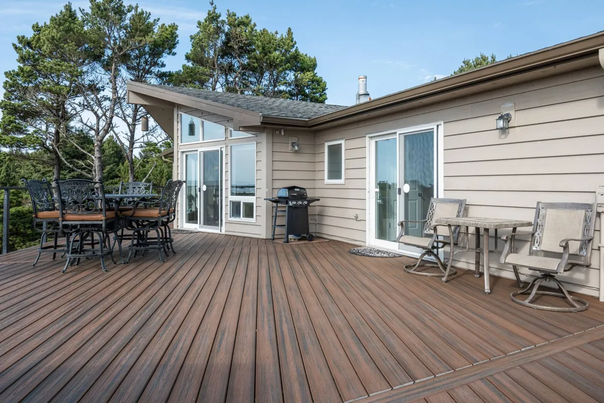 A large, inviting wooden deck surrounded by trees, featuring an outdoor dining table and a barbecue grill. The spacious area provides ample room for relaxation, alfresco meals, and play, with nature all around creating a tranquil setting.