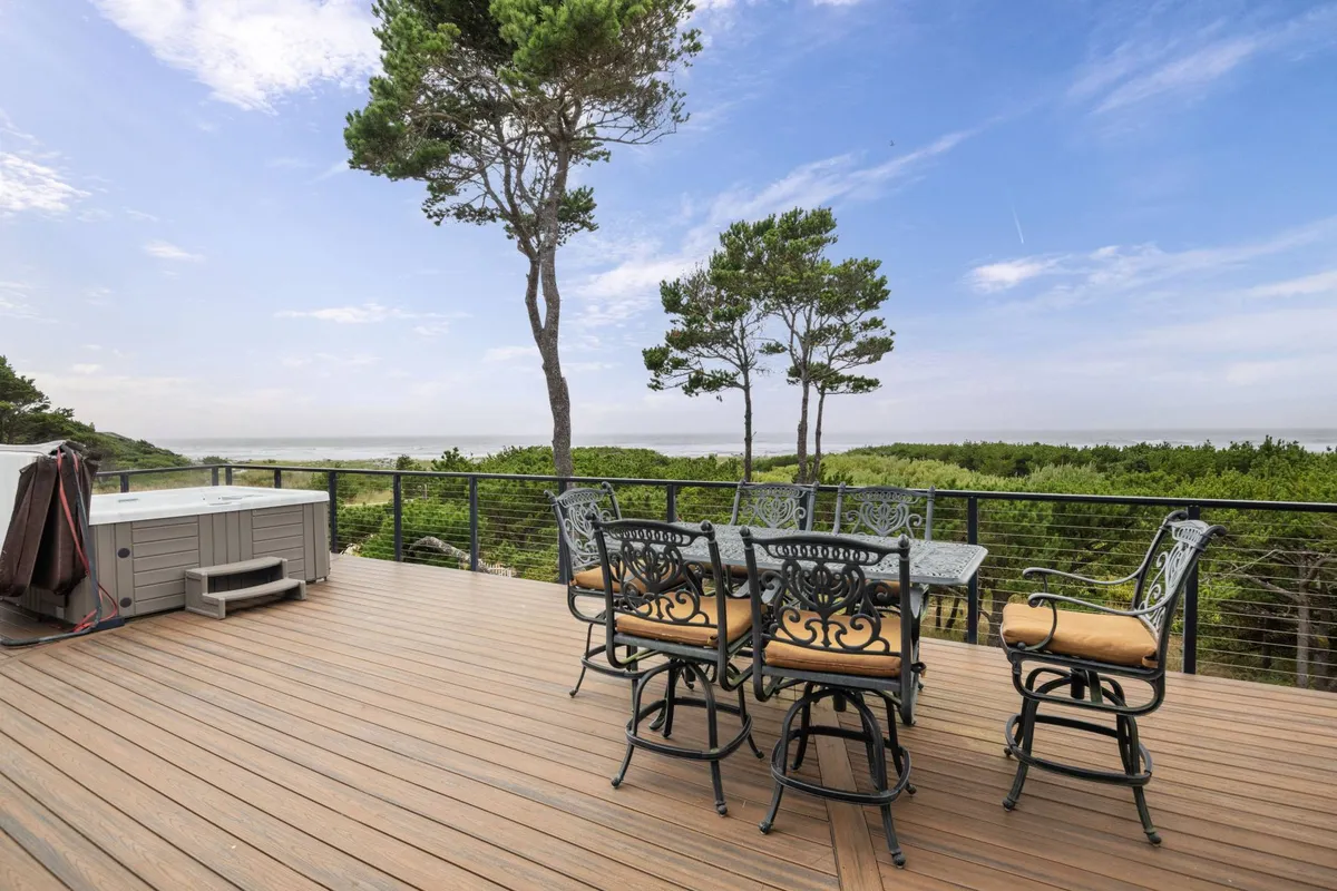 A spacious exterior deck featuring an outdoor dining set and a six-person hot tub, all with sweeping views of the ocean and surrounding landscape. The atmosphere is tranquil, perfect for relaxing, stargazing, or enjoying a meal outdoors while listening to the waves.