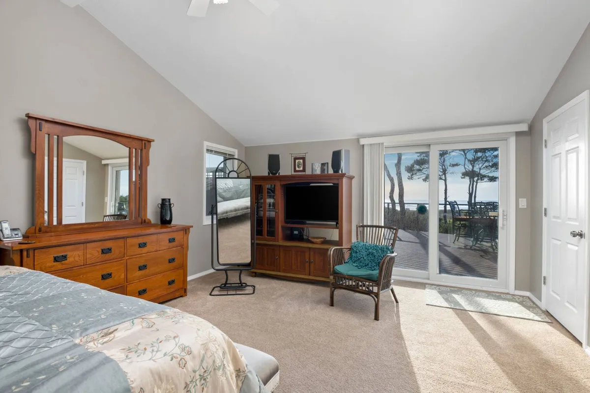 A cozy bedroom with patterned bedding on a queen bed, a stylish wooden dresser, and a television unit. Sliding glass doors open onto a deck, offering views and abundant natural light, while a comfortable chair and throw pillow complete the welcoming space.