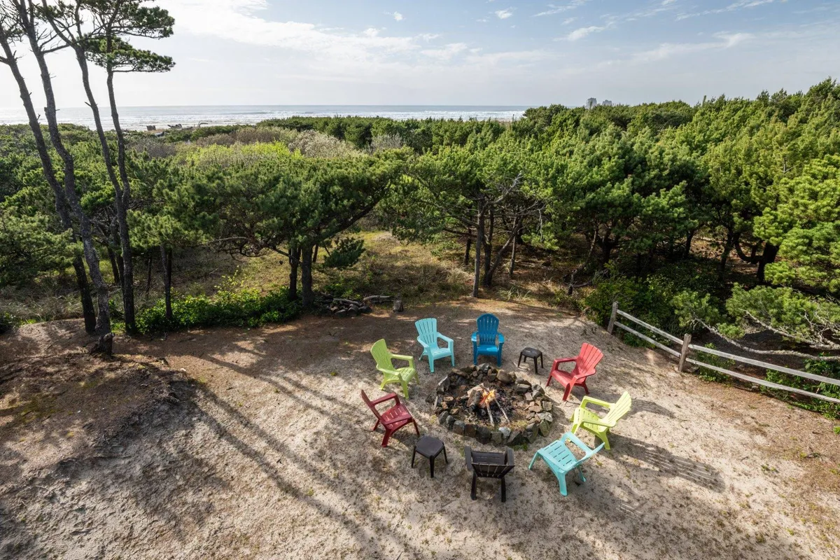 This exterior image features a fire pit surrounded by colorful chairs, nestled among lush trees with a stunning ocean view in the background. The setting exudes warmth and invites social gatherings or quiet moments in nature, tying directly to the section’s mention of relaxing by the fire-pit after a day of adventure.