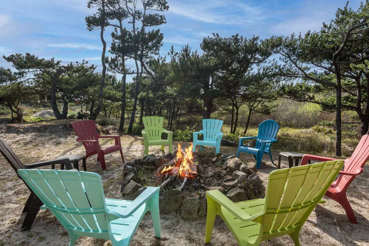 An inviting fire-pit area set outdoors among trees, surrounded by classic Adirondack chairs. The scene evokes warmth and togetherness, ideal for evening gatherings, storytelling, or stargazing under the open sky.