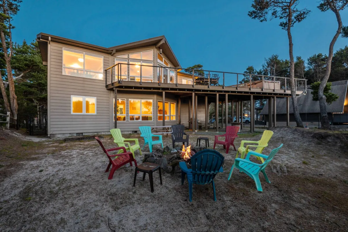 The image features a cozy fire pit surrounded by colorful chairs on the deck, with a view of the large house windows. The outdoor setting invites families to gather for stories, s'mores, and stargazing after a day’s adventure.